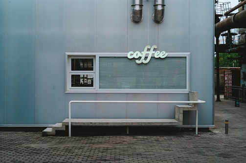 A minimalist coffee stand with a light blue metal exterior. The stand features a vintage-style 'coffee' sign in light green lettering above a window with closed blinds. A simplistic concrete bench and metal railing are positioned in front of the stand.