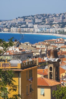 A coastal cityscape with a view of colorful residential buildings and a prominent clock tower in the foreground. The rooftops are predominantly red, and beyond the city lies a vast blue sea. In the background, a hillside is densely populated with more buildings, indicating a mountainous terrain.