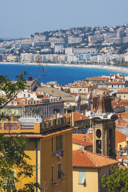 A coastal cityscape with a view of colorful residential buildings and a prominent clock tower in the foreground. The rooftops are predominantly red, and beyond the city lies a vast blue sea. In the background, a hillside is densely populated with more buildings, indicating a mountainous terrain.