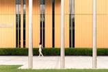 A woman walks along a paved sidewalk in front of a modern building with vertical orange panels and a row of windows. Three metallic poles stand in the forefront, and there is a well-maintained hedge lining the base of the building. The text 'FINANCIAL CENTER' is visible on the building.
