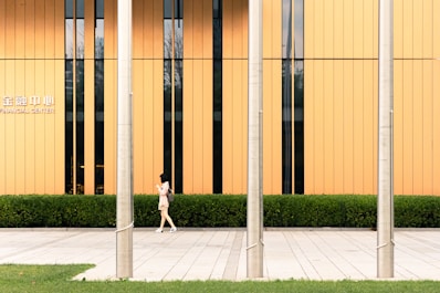 A woman walks along a paved sidewalk in front of a modern building with vertical orange panels and a row of windows. Three metallic poles stand in the forefront, and there is a well-maintained hedge lining the base of the building. The text 'FINANCIAL CENTER' is visible on the building.