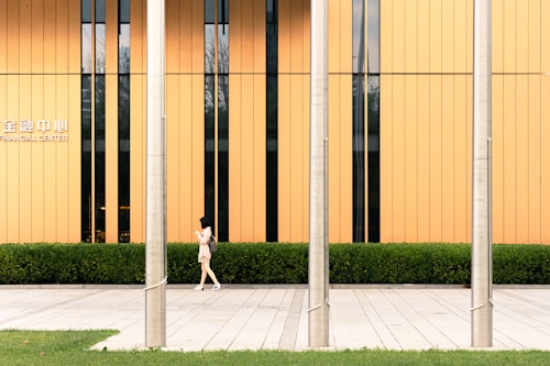 A woman walks along a paved sidewalk in front of a modern building with vertical orange panels and a row of windows. Three metallic poles stand in the forefront, and there is a well-maintained hedge lining the base of the building. The text 'FINANCIAL CENTER' is visible on the building.