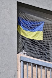 A blue and yellow flag is hanging behind a safety net on a balcony. The balcony is part of a building with a textured gray wall and vertical railing bars.