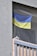 A blue and yellow flag is hanging behind a safety net on a balcony. The balcony is part of a building with a textured gray wall and vertical railing bars.