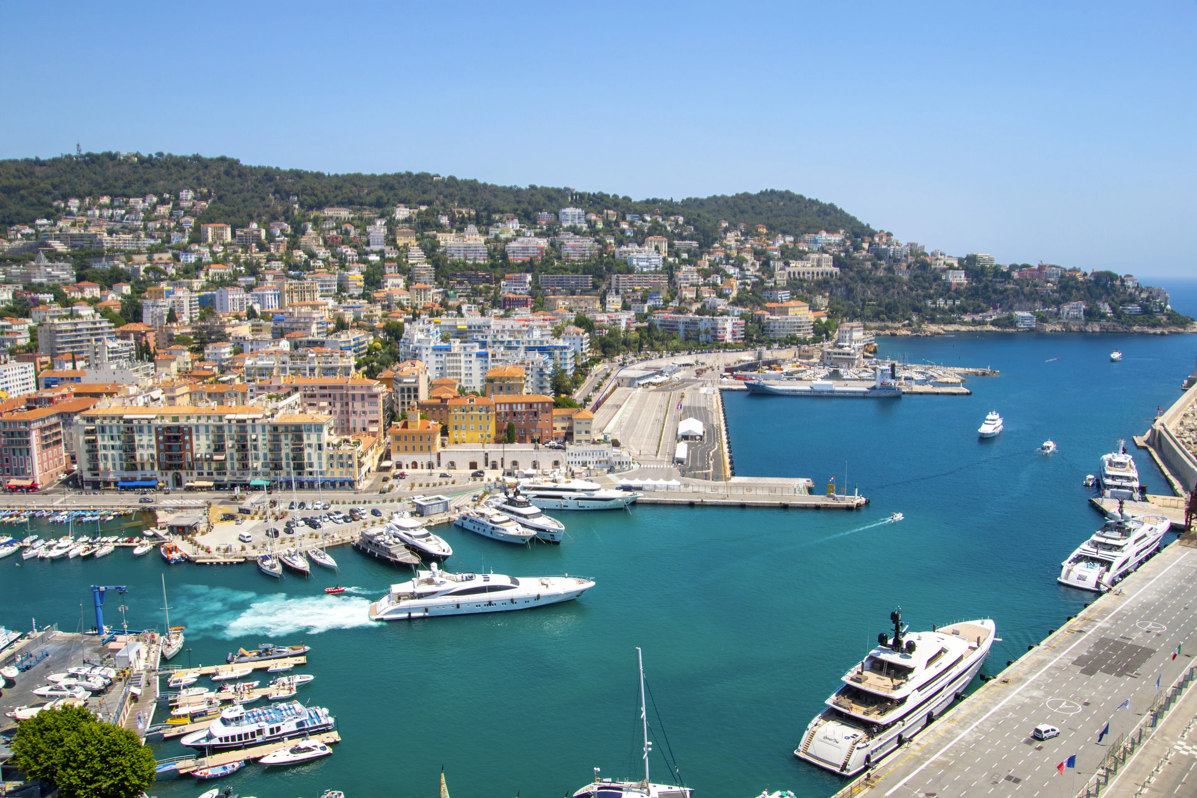 Port Lympia in Nice packed with yachts and motorboats, pastel apartment blocks along the quay and a green hillside rising behind.
