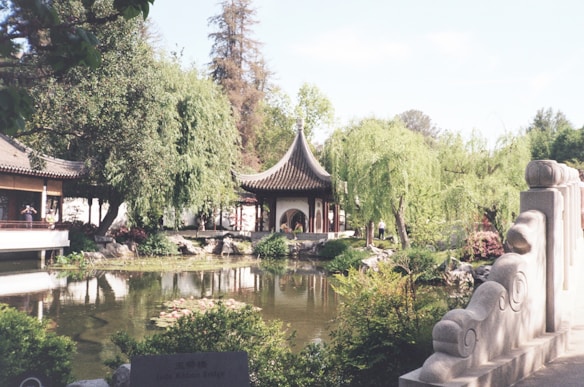 A serene garden scene features a traditional Chinese architecture pavilion set amidst lush greenery, including tall willow trees. The area is reflected in a tranquil pond adorned with water lilies. Decorative stone railings enhance the peaceful ambiance, and a few people can be seen enjoying the scenery.