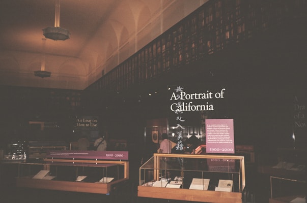 The image depicts an interior of a library or exhibition space with dim lighting. The foreground features display cases with books or documents, labeled with years such as 1900-2000. There's a prominent sign titled 'A Portrait of California' with additional text panels. The ceiling is high with decorative light fixtures, and the walls are lined with bookshelves.