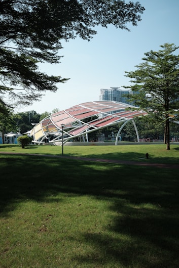 A modern architectural structure with an arched, multicolored canopy is situated in a park. Surrounding the structure is a well-maintained lawn with scattered trees providing shade. In the background, a tall building and more greenery are visible.