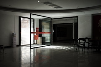 A dimly lit indoor space featuring a glass sliding door leading to another room. The light from outside creates reflections on the glossy floor. Several empty chairs are arranged near the door, and there is a notice board with red signage partially visible. The atmosphere is quiet and somewhat stark.