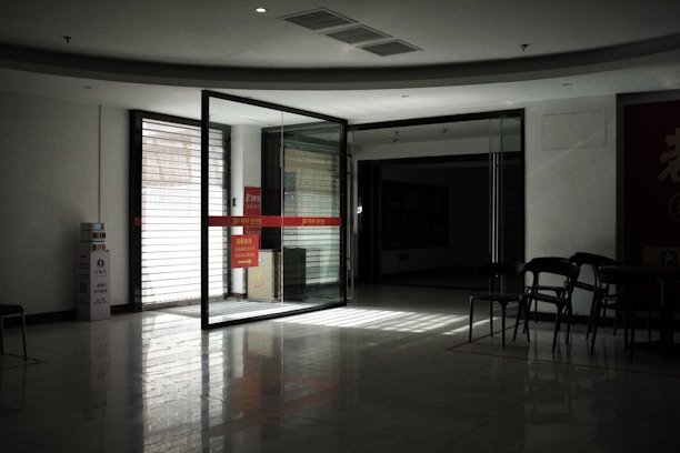 A dimly lit indoor space featuring a glass sliding door leading to another room. The light from outside creates reflections on the glossy floor. Several empty chairs are arranged near the door, and there is a notice board with red signage partially visible. The atmosphere is quiet and somewhat stark.