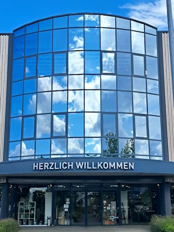 The image shows the glass facade of a modern building reflecting a bright blue sky with white clouds. At the bottom, there is an entryway with the sign 'HERZLICH WILLKOMMEN' written above it. The view includes reflections of trees as well as interior elements visible through the glass doors.