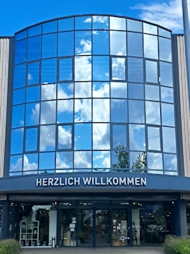 The image shows the glass facade of a modern building reflecting a bright blue sky with white clouds. At the bottom, there is an entryway with the sign 'HERZLICH WILLKOMMEN' written above it. The view includes reflections of trees as well as interior elements visible through the glass doors.