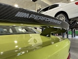 A close-up view of a car's rear spoiler with 'M Performance' branding, situated in a garage or workshop. The car is lime green in color. In the background, a white car is elevated on a hydraulic lift.