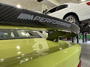 A close-up view of a car's rear spoiler with 'M Performance' branding, situated in a garage or workshop. The car is lime green in color. In the background, a white car is elevated on a hydraulic lift.