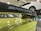 A close-up view of a car's rear spoiler with 'M Performance' branding, situated in a garage or workshop. The car is lime green in color. In the background, a white car is elevated on a hydraulic lift.
