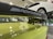 A close-up view of a car's rear spoiler with 'M Performance' branding, situated in a garage or workshop. The car is lime green in color. In the background, a white car is elevated on a hydraulic lift.