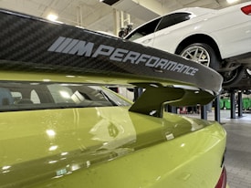 A close-up view of a car's rear spoiler with 'M Performance' branding, situated in a garage or workshop. The car is lime green in color. In the background, a white car is elevated on a hydraulic lift.