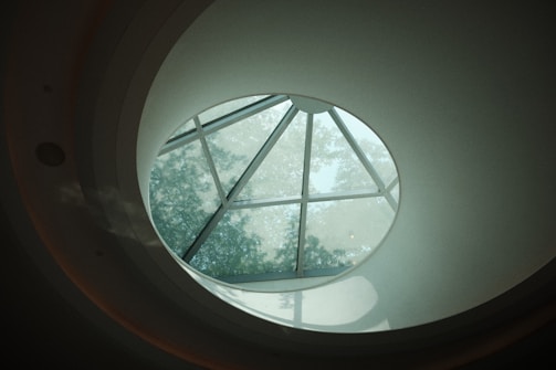 Circular skylight with multiple glass panels set within a domed ceiling, allowing soft natural light and tree silhouettes to filter through.