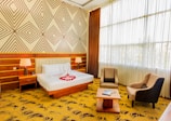 A well-lit hotel room featuring a large double bed with white linens and red rose petals arranged in a heart shape. The bed has a headboard and is flanked by two bedside tables with modern lamps. Wooden paneling and geometric patterned wallpaper adorn the walls. Two upholstered armchairs and a coffee table are set near a large window with sheer curtains, allowing plenty of natural light to pour in.