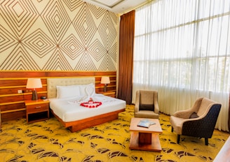 A well-lit hotel room featuring a large double bed with white linens and red rose petals arranged in a heart shape. The bed has a headboard and is flanked by two bedside tables with modern lamps. Wooden paneling and geometric patterned wallpaper adorn the walls. Two upholstered armchairs and a coffee table are set near a large window with sheer curtains, allowing plenty of natural light to pour in.