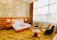 A well-lit hotel room featuring a large double bed with white linens and red rose petals arranged in a heart shape. The bed has a headboard and is flanked by two bedside tables with modern lamps. Wooden paneling and geometric patterned wallpaper adorn the walls. Two upholstered armchairs and a coffee table are set near a large window with sheer curtains, allowing plenty of natural light to pour in.