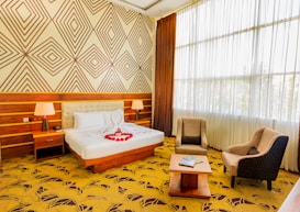 A well-lit hotel room featuring a large double bed with white linens and red rose petals arranged in a heart shape. The bed has a headboard and is flanked by two bedside tables with modern lamps. Wooden paneling and geometric patterned wallpaper adorn the walls. Two upholstered armchairs and a coffee table are set near a large window with sheer curtains, allowing plenty of natural light to pour in.