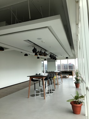 A modern, open office space with long tables and high stools arranged neatly. There are black pendant lights hanging from the ceiling. The area has large windows that let in natural light, with a view of buildings outside. Potted plants add a touch of greenery to the otherwise minimalist decor.