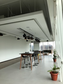 A modern, open office space with long tables and high stools arranged neatly. There are black pendant lights hanging from the ceiling. The area has large windows that let in natural light, with a view of buildings outside. Potted plants add a touch of greenery to the otherwise minimalist decor.