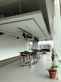 A modern, open office space with long tables and high stools arranged neatly. There are black pendant lights hanging from the ceiling. The area has large windows that let in natural light, with a view of buildings outside. Potted plants add a touch of greenery to the otherwise minimalist decor.