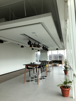 A modern, open office space with long tables and high stools arranged neatly. There are black pendant lights hanging from the ceiling. The area has large windows that let in natural light, with a view of buildings outside. Potted plants add a touch of greenery to the otherwise minimalist decor.