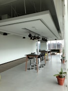 A spacious modern office area with a high ceiling, featuring a large table surrounded by stools and potted plants placed along the perimeter. Pendant lights hang from the ceiling, and natural light floods the room through floor-to-ceiling windows. In the background, an urban landscape is visible through the windows.