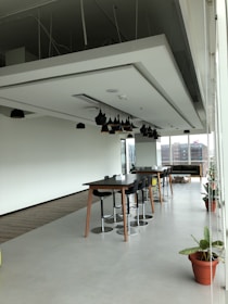 A spacious modern office area with a high ceiling, featuring a large table surrounded by stools and potted plants placed along the perimeter. Pendant lights hang from the ceiling, and natural light floods the room through floor-to-ceiling windows. In the background, an urban landscape is visible through the windows.