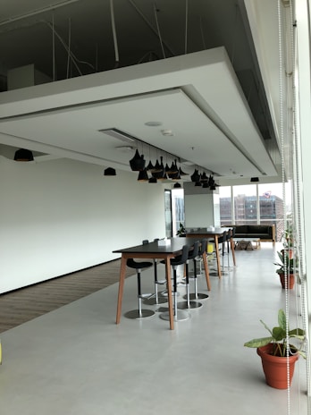 A spacious modern office area with a high ceiling, featuring a large table surrounded by stools and potted plants placed along the perimeter. Pendant lights hang from the ceiling, and natural light floods the room through floor-to-ceiling windows. In the background, an urban landscape is visible through the windows.