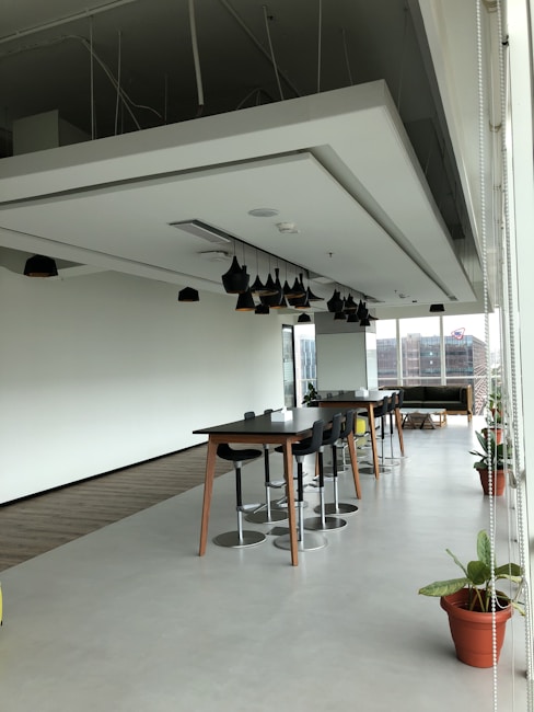 A spacious modern office area with a high ceiling, featuring a large table surrounded by stools and potted plants placed along the perimeter. Pendant lights hang from the ceiling, and natural light floods the room through floor-to-ceiling windows. In the background, an urban landscape is visible through the windows.
