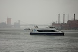 A ferry travels on a river with an industrial skyline in the background, including a bridge and several tall smokestacks. The water appears calm and the sky is overcast, suggesting a gloomy or rainy day.