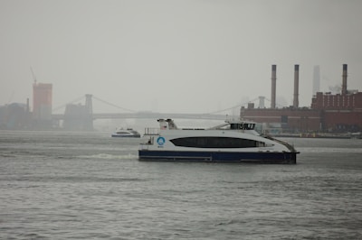 A ferry travels on a river with an industrial skyline in the background, including a bridge and several tall smokestacks. The water appears calm and the sky is overcast, suggesting a gloomy or rainy day.