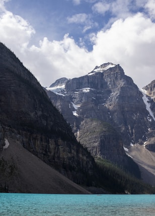 Majestic mountain range with rugged cliffs covered in patches of snow and surrounded by a forested landscape. Below, a vibrant turquoise lake reflects the natural beauty of the scenery. The sky is partly cloudy, adding to the serene and untouched wilderness atmosphere.