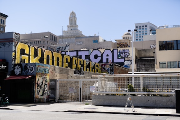 A vibrant urban scene featuring large graffiti art on a wall, with various city buildings in the background. The graffiti is bold, with large letters and intricate details, and is surrounded by other smaller pieces of street art. The architecture in the background adds contrast to the colorful graffiti. The scene conveys a feeling of urban culture and expression.