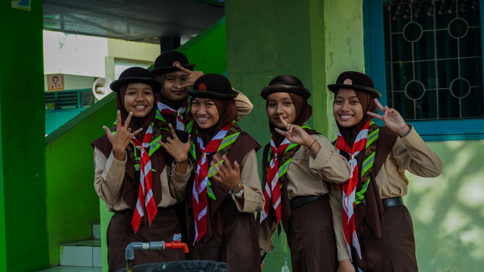 A group of five young individuals wearing scout uniforms stand together, smiling and showing peace signs with their fingers. The uniforms consist of brown hats, beige shirts, and red and white scarves. The background features a green wall and a window with bars, along with some stairs.