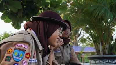 Two individuals wearing scout uniforms are sitting outside surrounded by greenery. One person is smiling, wearing a maroon hat and glasses, while the other looks on thoughtfully. Various colorful patches adorn their uniforms, indicating affiliations or achievements. Palm trees and a large leafy plant are in the background, creating a tropical atmosphere.