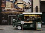 A green vintage coffee truck is parked on a city street. The truck has a window displaying various coffee items for sale and a sign promoting 'Ralph's Coffee.' There is an elegant storefront in the background with large glass windows and an advertisement for luxury brands, creating a blend of classic and modern urban aesthetics.