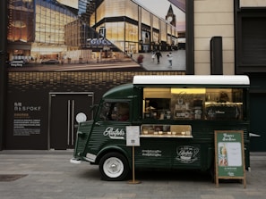 A green vintage coffee truck is parked on a city street. The truck has a window displaying various coffee items for sale and a sign promoting 'Ralph's Coffee.' There is an elegant storefront in the background with large glass windows and an advertisement for luxury brands, creating a blend of classic and modern urban aesthetics.