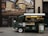 A green vintage coffee truck is parked on a city street. The truck has a window displaying various coffee items for sale and a sign promoting 'Ralph's Coffee.' There is an elegant storefront in the background with large glass windows and an advertisement for luxury brands, creating a blend of classic and modern urban aesthetics.