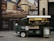 A green vintage coffee truck is parked on a city street. The truck has a window displaying various coffee items for sale and a sign promoting 'Ralph's Coffee.' There is an elegant storefront in the background with large glass windows and an advertisement for luxury brands, creating a blend of classic and modern urban aesthetics.