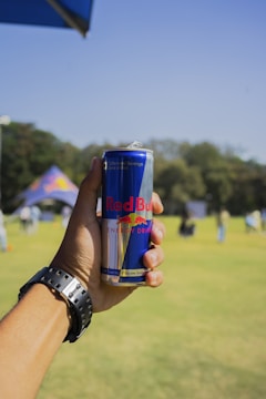 A hand holding a can of Red Bull energy drink with a backdrop of a grassy field and trees. The person wearing a watch is in focus, while the background is slightly blurred.