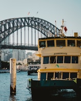 A ferry named Scarborough is docked in the foreground with a visible Transport Sydney Ferries logo. The iconic Sydney Harbour Bridge looms in the background, with a cityscape of tall buildings partially obscured by the bridge's steel structure. Two flags are flying at the top of the bridge, and another ferry is seen in the midground navigating through the water.