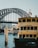 A ferry named Scarborough is docked in the foreground with a visible Transport Sydney Ferries logo. The iconic Sydney Harbour Bridge looms in the background, with a cityscape of tall buildings partially obscured by the bridge's steel structure. Two flags are flying at the top of the bridge, and another ferry is seen in the midground navigating through the water.