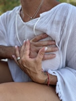 Hands gently rest on a person's chest, creating a sense of comfort and connection. The person is wearing a white textured garment, and there's a red bracelet on one of the hands. The scene is intimate, emphasizing human touch and emotion.