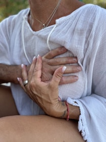 Hands gently rest on a person's chest, creating a sense of comfort and connection. The person is wearing a white textured garment, and there's a red bracelet on one of the hands. The scene is intimate, emphasizing human touch and emotion.