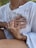 Hands gently rest on a person's chest, creating a sense of comfort and connection. The person is wearing a white textured garment, and there's a red bracelet on one of the hands. The scene is intimate, emphasizing human touch and emotion.